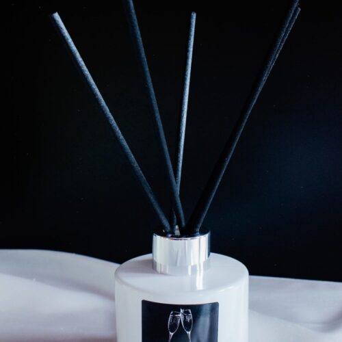 Dyfuzor zapachowy o zapachu prosecco w białej butelce ze srebrną zakrętką i czarnymi patyczkami.