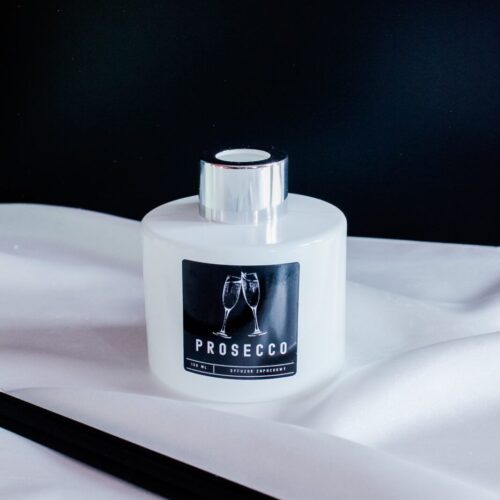 Dyfuzor zapachowy o zapachu prosecco w białej butelce ze srebrną zakrętką i czarnymi patyczkami.